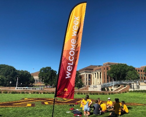 A maroon and gold Welcome Week flag on the Univeristy of Minnesota lawn, with students sitting on the grass